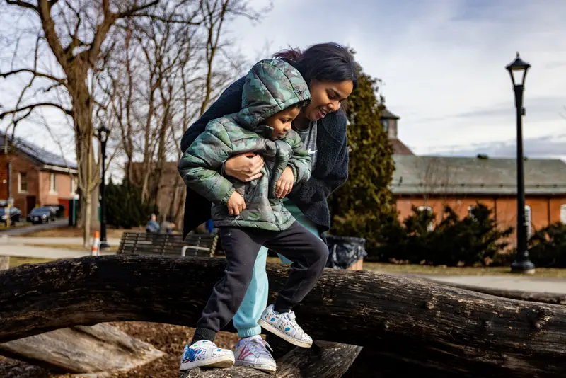 A smiling woman helps a small boy hop between logs.