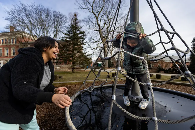 A smiling woman reaches toward a piece of playground equipment that a small boy is standing on while holding onto rope netting.