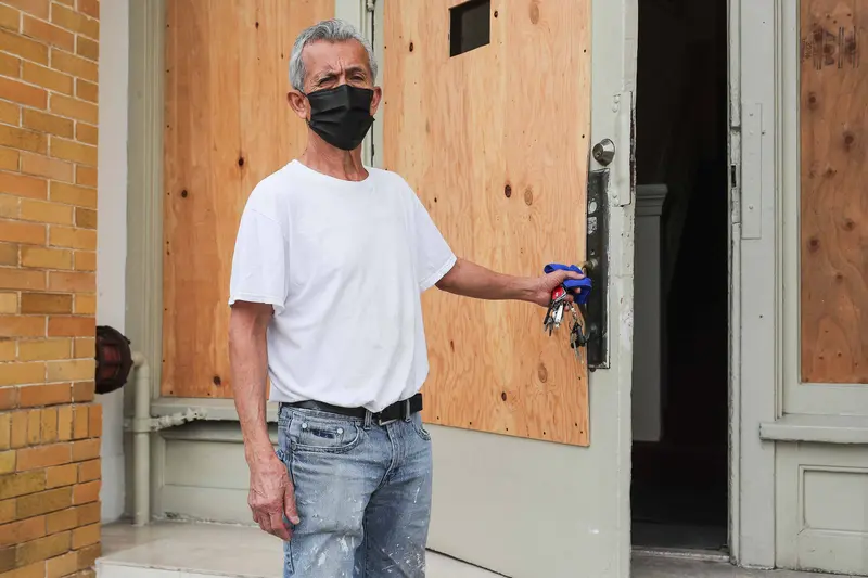 A man with grey hair wearing a black surgical mask opens a boarded up door into a hotel lobby.