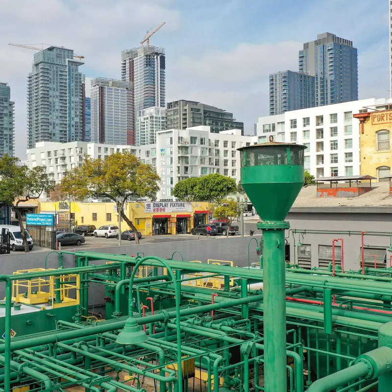 A petroleum well operation consisting of a network of green pipes, sits in the foreground of the Los Angleles downtown cityscape.