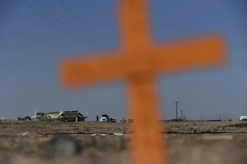 An out-of-focus cross planted in the dirt in the foreground with cleanup crews working around vehicles in the distant background.