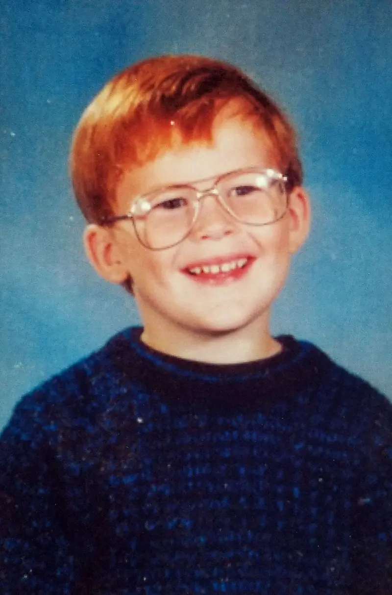 A small boy in glasses and a blue sweater smiles in a school photo