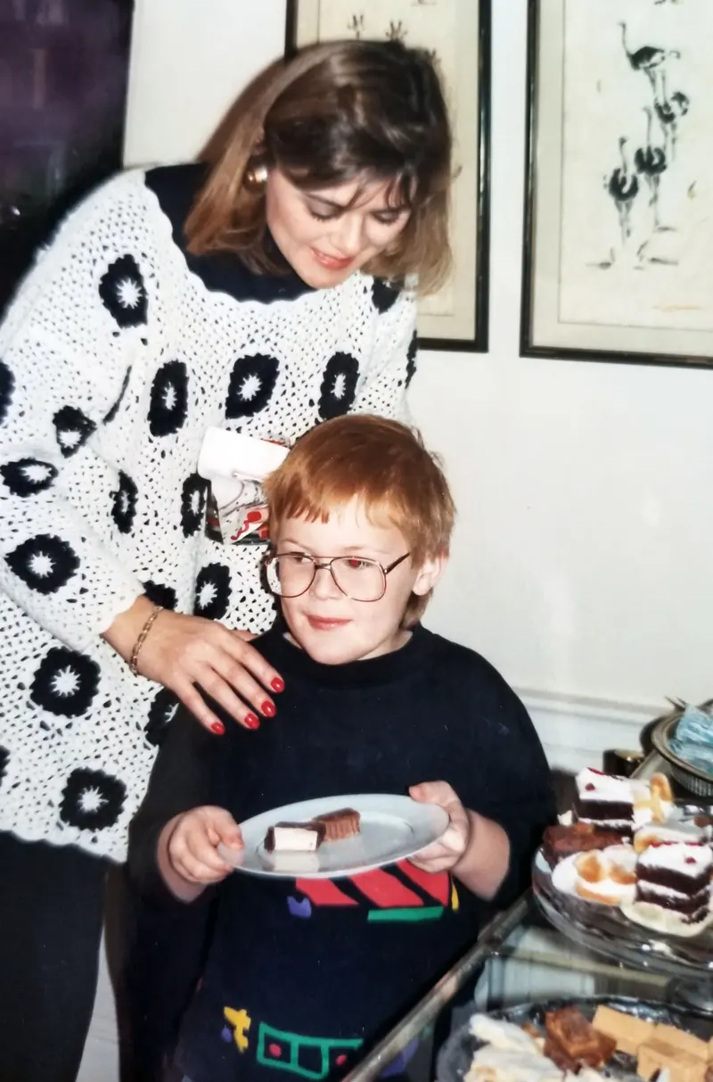 A woman leans over a small boy in glasses who's holding a plate of cookies.