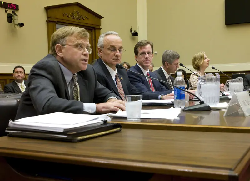 Four men and a women in businesswear sit on one side of a long table covered in microphones and papers.