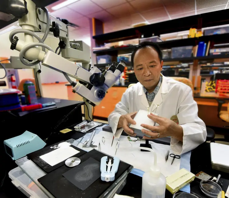 Tsien at a workbench wearing a lab coat and surrounded by instruments and a microscope eyepiece.
