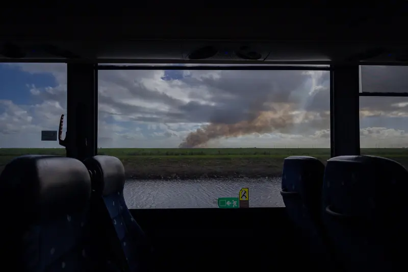 Fields viewed through a bus window. In the center of the frame, a plume of smoke rises.