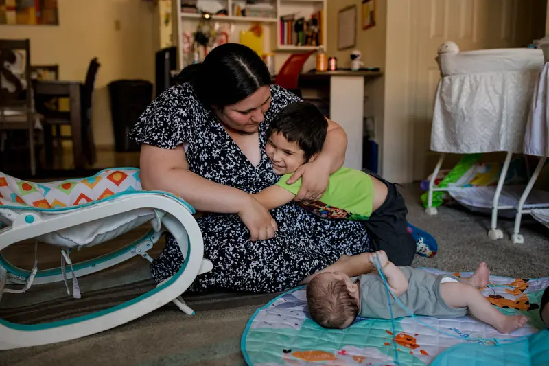 A woman sitting on a carpeted floor hugs a toddler with one arm while a baby lies on their back on a play mat.