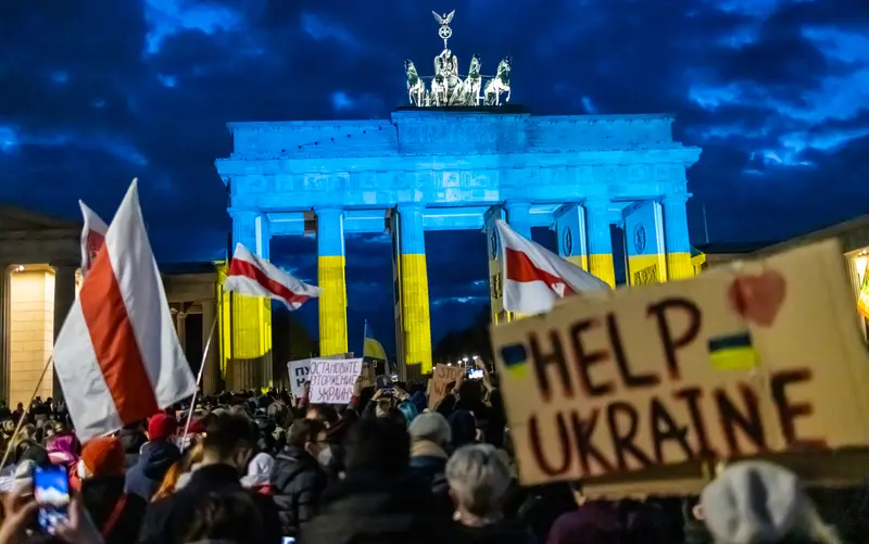 A crowd of people with signs and flags in front of a huge columned gate that's lit up in blue over yellow, like the Ukrainian flag.
