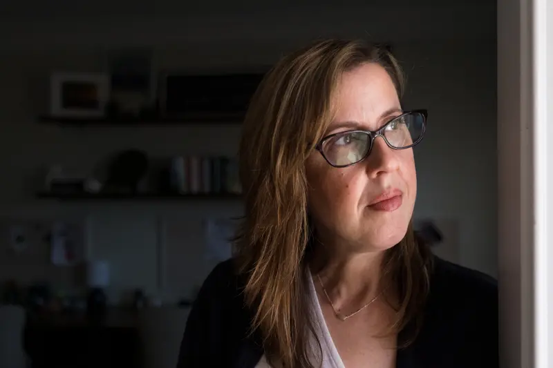 A white woman with straight brown hair and glasses off to the right, where light from a window is coming in.