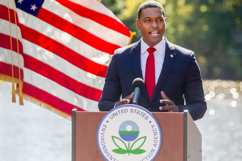 A Black man in a suit stands at a lectern bearing the EPA logo. An American flag billows behind him on the left, and water sparkles in the background.