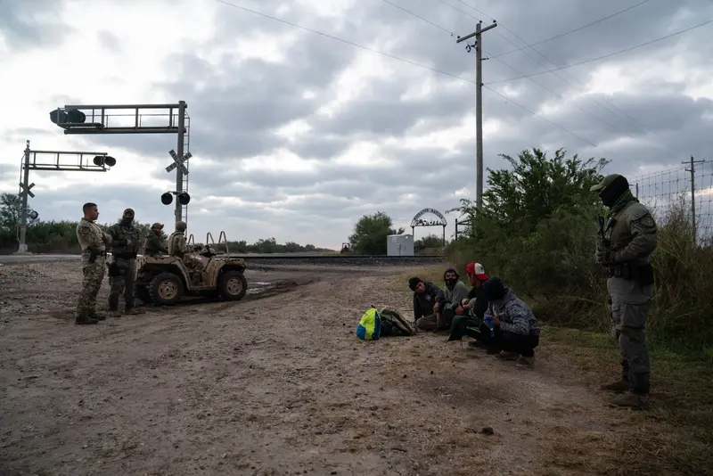 Four men sit or squat on the ground while five armed men in camouflage gather around them. In the background are train tracks and a tall, arched gate.