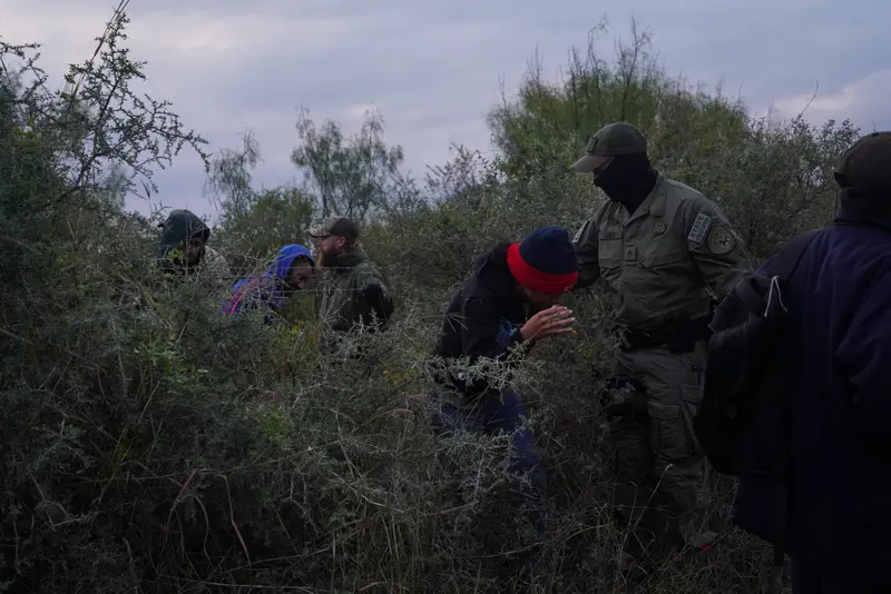 People duck through some tall brush while uniformed agents look on and direct them where to go.
