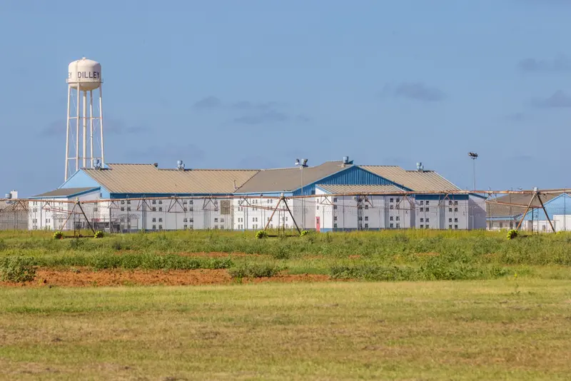 An open field runs up to a rusty fence, beyond which is a white, two-story building with blue accents. Beside the building is a water tower that says Dilley.