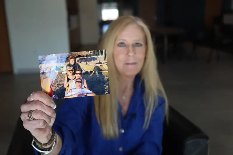 A blonde woman in a blue shirt holds a photo of four people posing together at the beach.