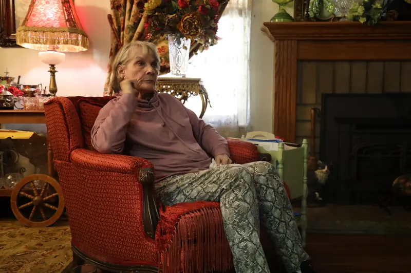 An older woman sits in an upholstered armchair in a room full of knickknacks.