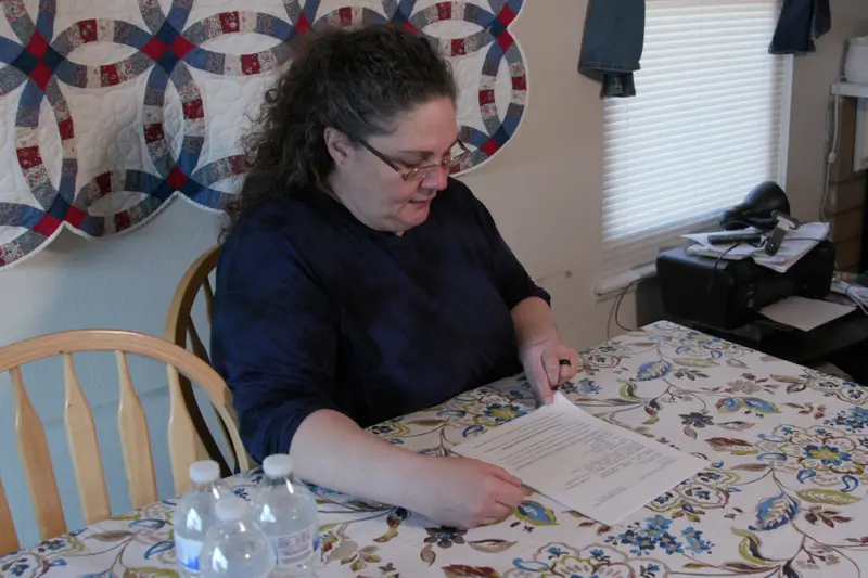 A woman with dark curly hair reads a document at a kitchen table. A quilt hangs on the wall behind her.