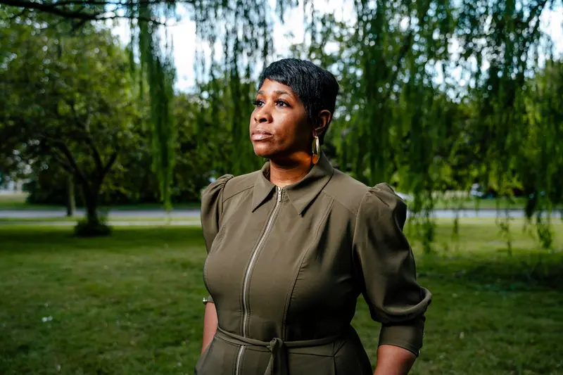 A woman in a dark olive dress stands in front of a tree.