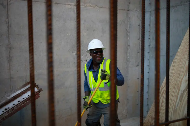 A Black man in a hard hat, gloves and yellow safety vest laughs while holding a long-handled tool. He is surrounded by concrete walls and upright pieces of rebar.