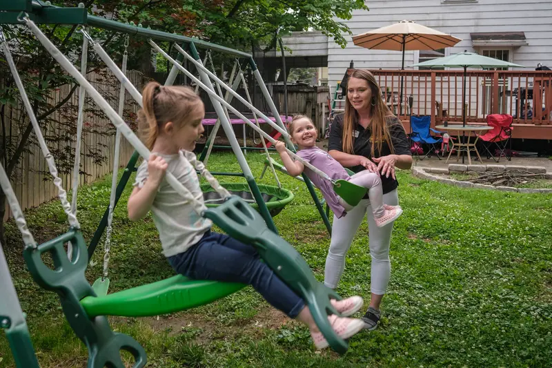 Two girls swing on a swing set in a grassy, fenced-in backyard while a white woman pushes the smaller girl.