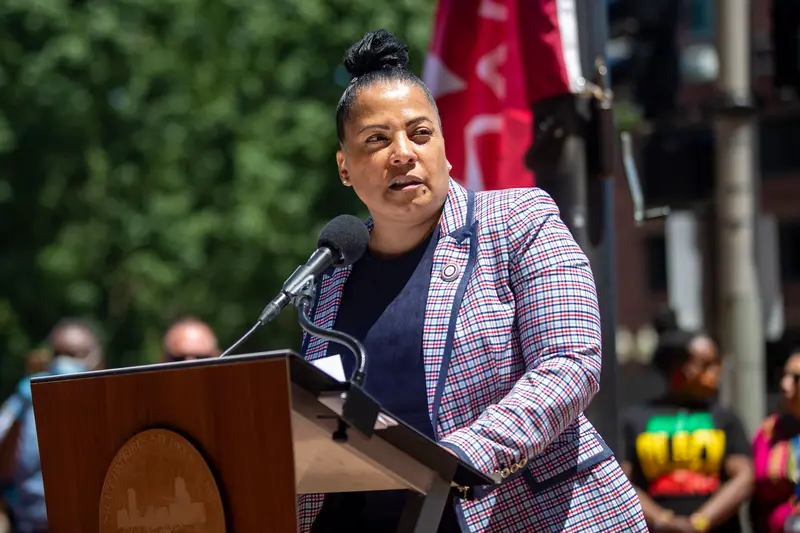 Rachael Rollins, a Black woman in a plaid suit, speaks at a microphone outdoors.