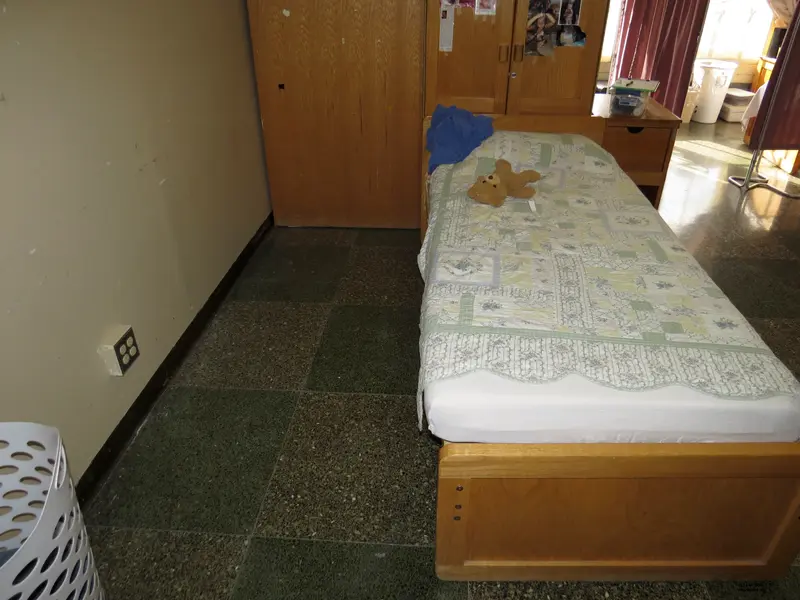 A twin bed with a teddy bear and a quilt on it. It's in an institutional-style room with beige walls and wooden storage furniture.