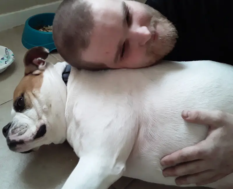 A white man with short hair and a blond beard lies on the floor with his head on a dog's shoulder and his hand on its belly.