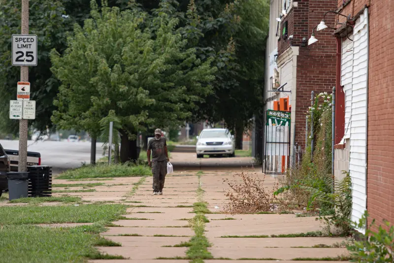 A man walks down a sidewalk with plants growing between the cracks.