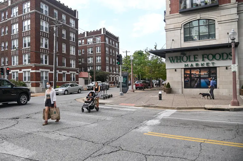 One woman with shopping bags and one with a stroller cross the street in front of a Whole Foods.