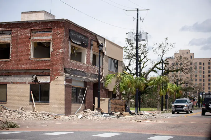 A brick building with nearly all its windows broken and huge chunks of its facade missing. Debris blocks the sidewalk and parts of the street around the building.
