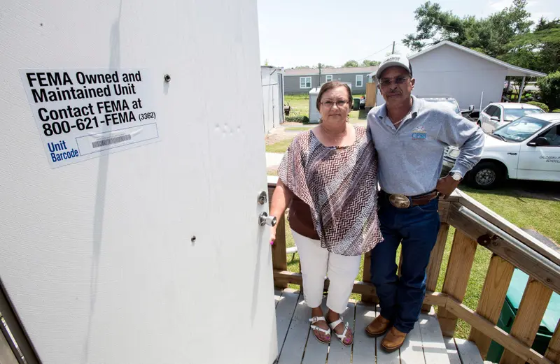 A couple stand outside on wooden steps next to a door with a sign that says "FEMA owned and maintained unit."