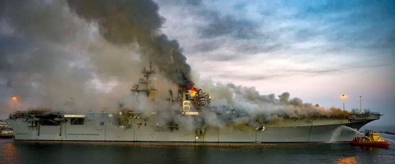 Smoke pours out of a Navy ship as a nearby fireboat applies a spray of water.