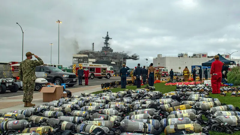Military officers salute in the direction of a burning ship. Nearby, a large pile of gas cylinders rests on a lawn.
