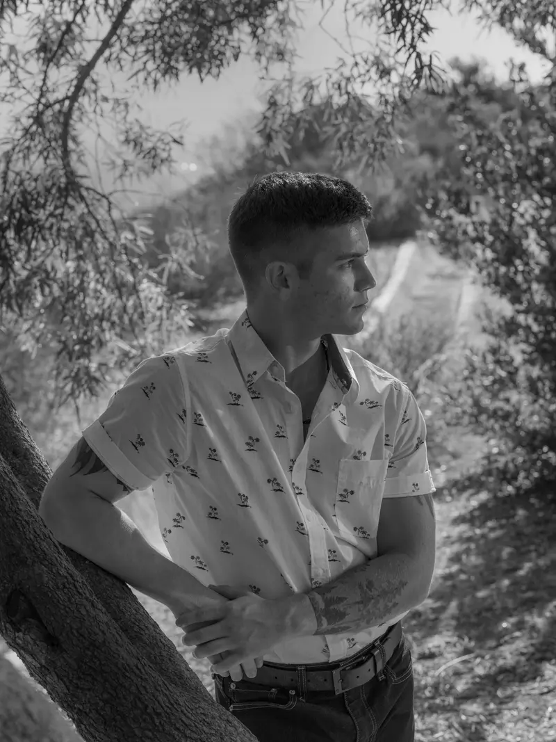 A young man in a white patterned shirt leans against a tree.