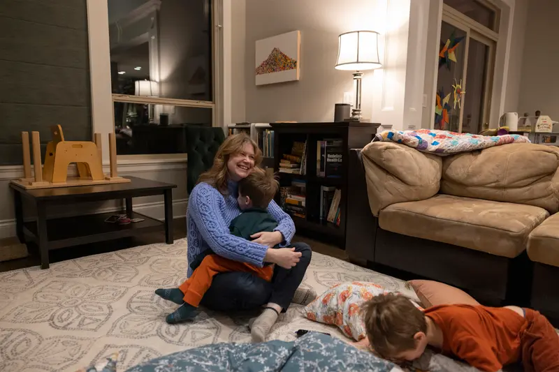 The woman hugs one boy while another lies on cushions on the floor nearby.