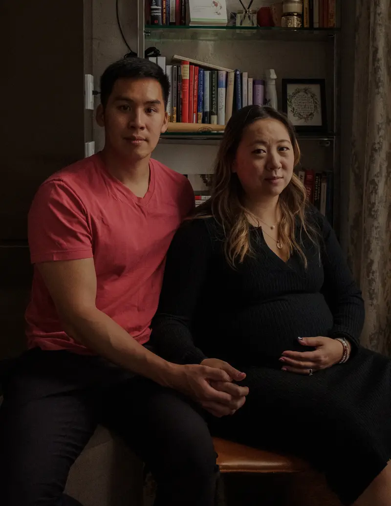 A pregnant woman holds her belly while sitting next to her husband in front of a bookshelf.