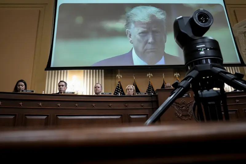 Donald Trump's face appears on a large video screen behind a row of Congressional committee members.