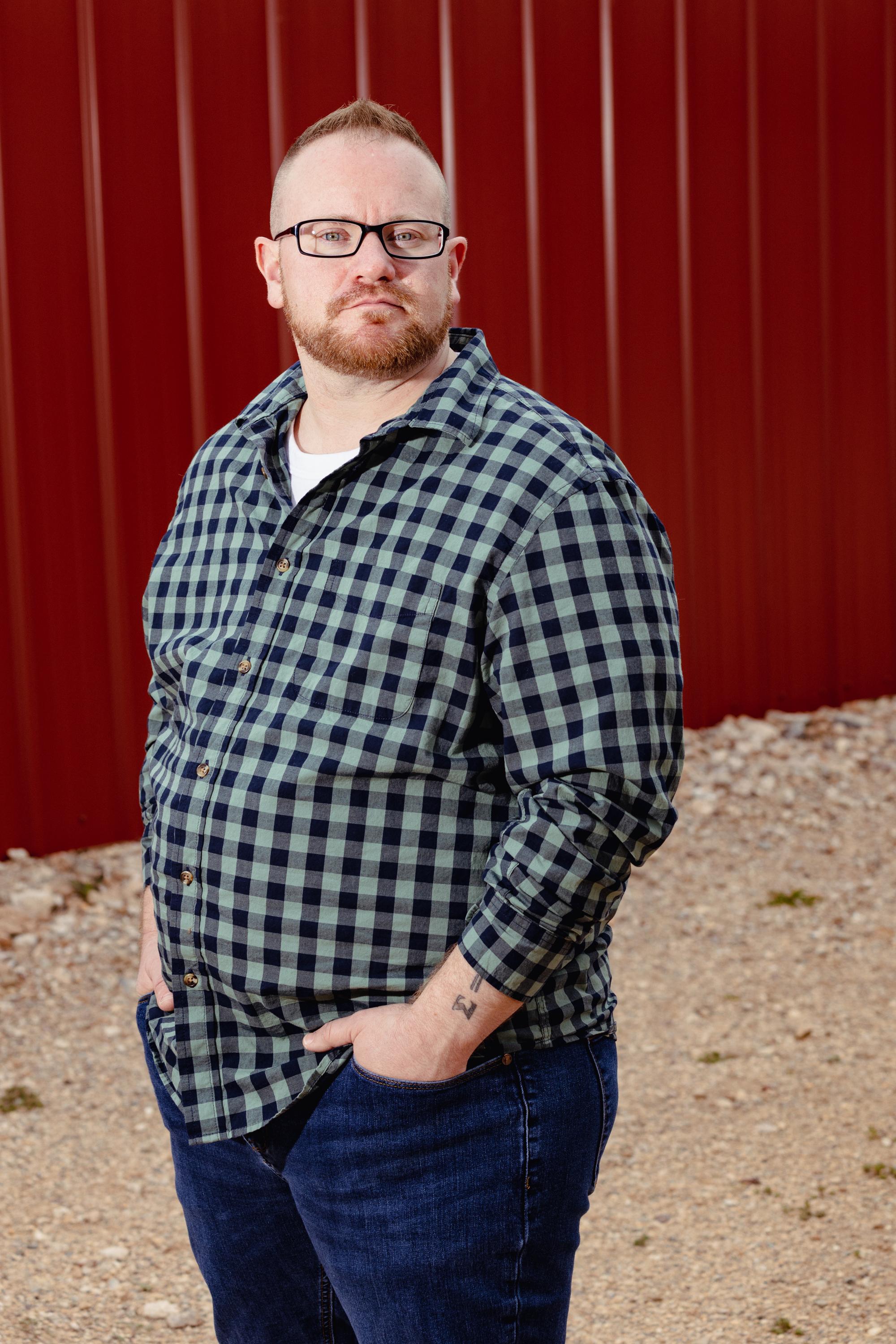 A man with reddish hair and a beard wears a green checked shirt, jeans, and rectangular glasses. He’s standing in a dirt area in front of a red wall.