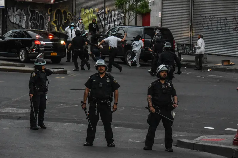 NYPD officers in riot gear stand along a Bronx street; in the background, two officers wield batons as civilians flee.