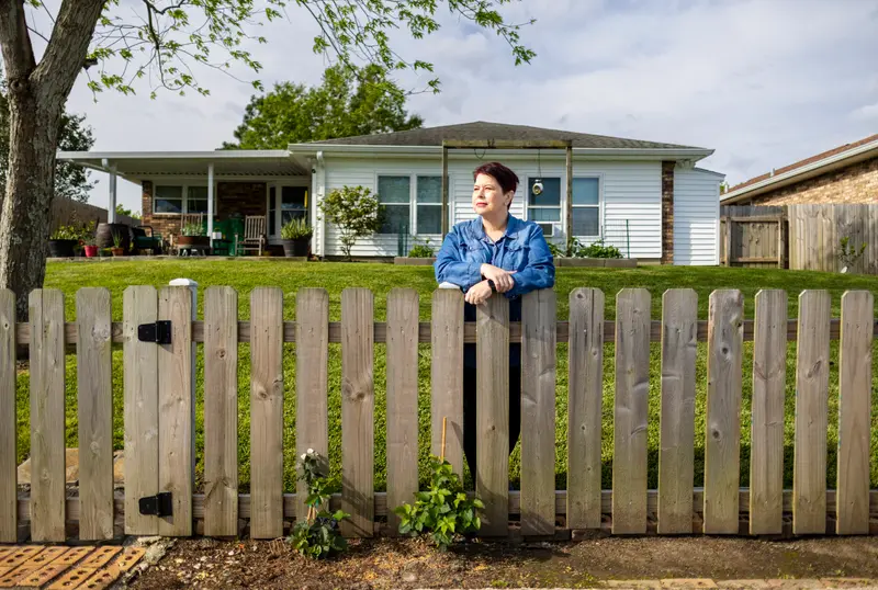 A woman in a denim shirt stands behind a wood fence surrounding a one-story home.
