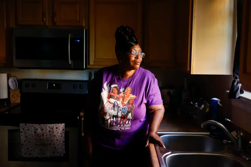 A woman wearing glasses and a purple memorial t-shirt stands in a kitchen.