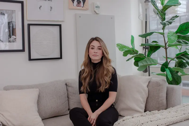 A white woman dressed all in black on a tan couch, her blonde hair in loose waves.