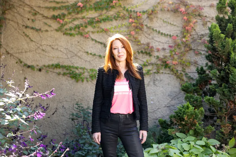 A white woman with red hair stands outside. She's wearing dark jeans, a blazer and a pink T-shirt.