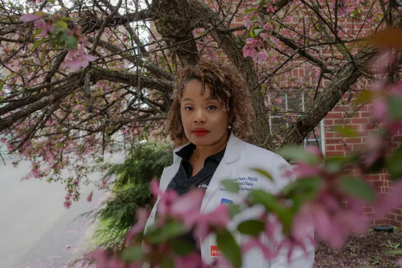 A Black woman in a lab coat stands amidst the branches of a tree with pink blossoms.