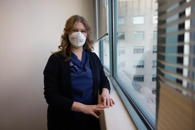 A white woman in a face mask and scrubs stands by a window.