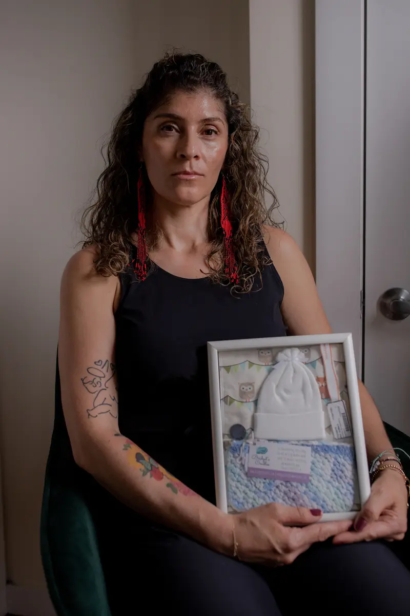 A woman with curly hair holds a frame containing a baby's hat, a blanket, a hospital wristband and other baby items.