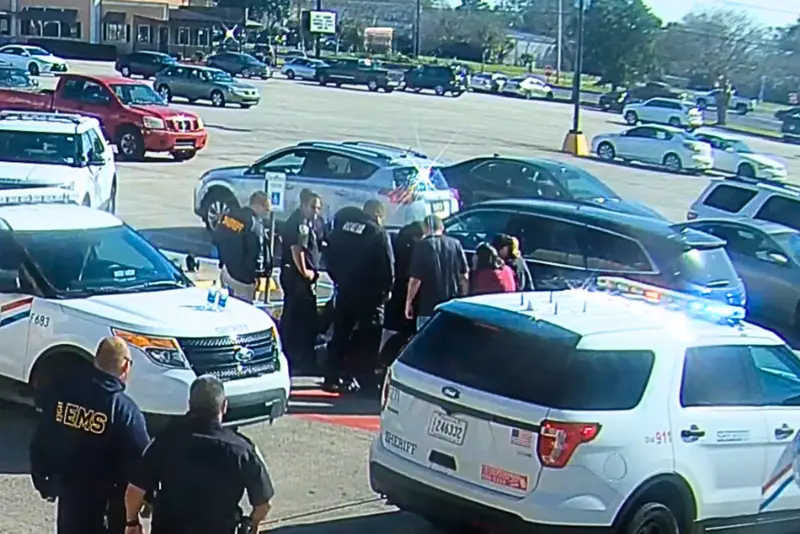 A crowd of police officers and EMTs stand around someone on the ground in a parking lot.