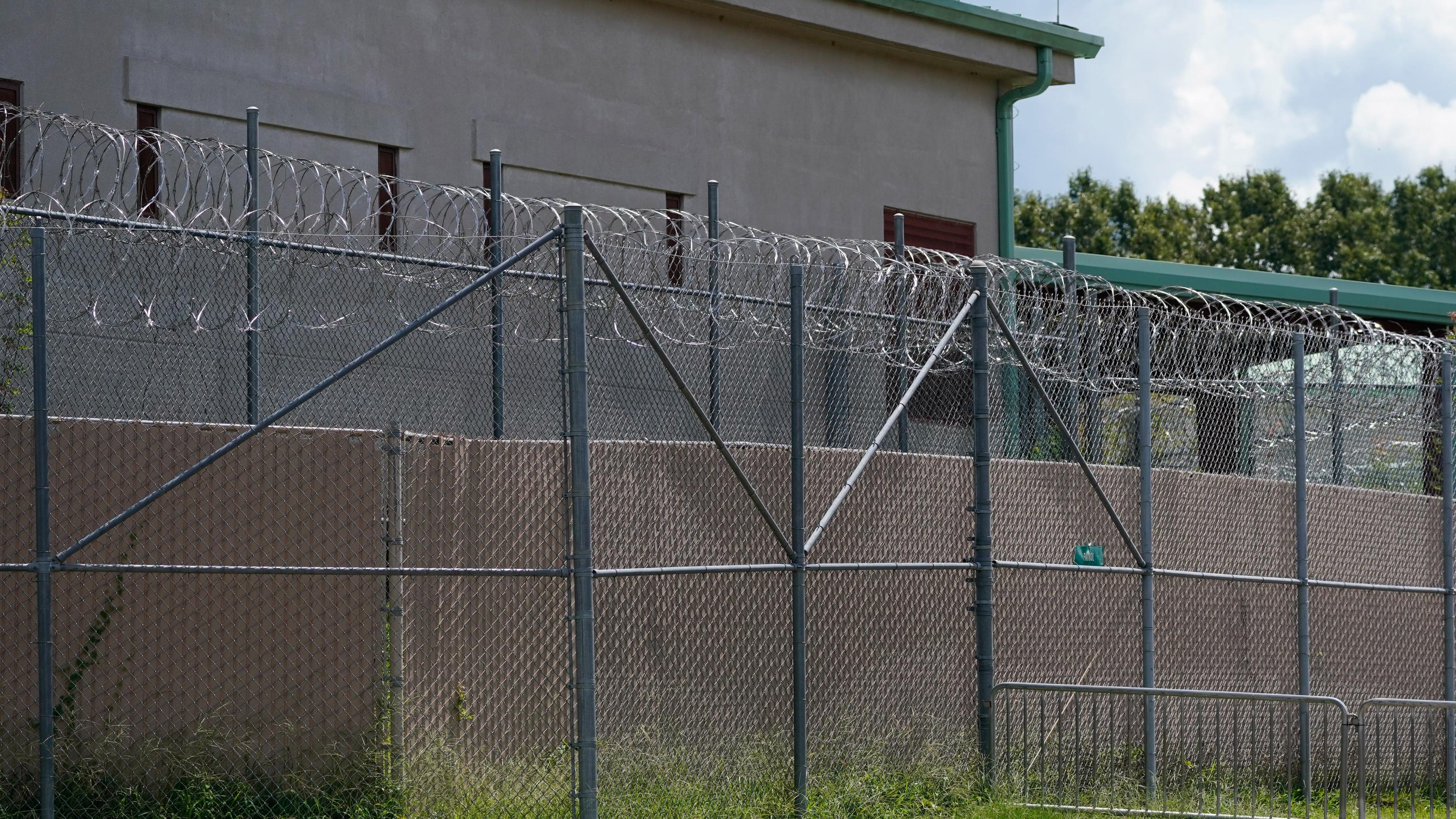 A gray detention center with pale green gutters, behind a barbed wire fence.