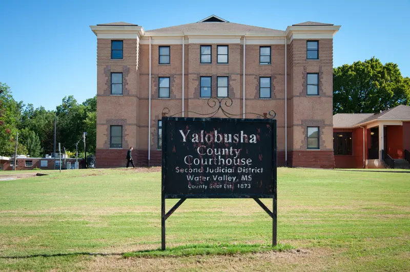 A black, rusted sign reads “Yalobusha County Courthouse” in front of a brick building.