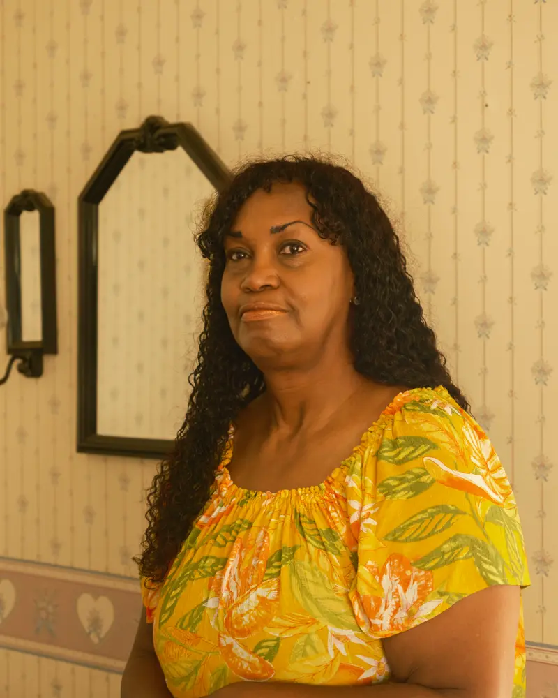 An African American woman in a flowered top stands leaning against a door frame.
