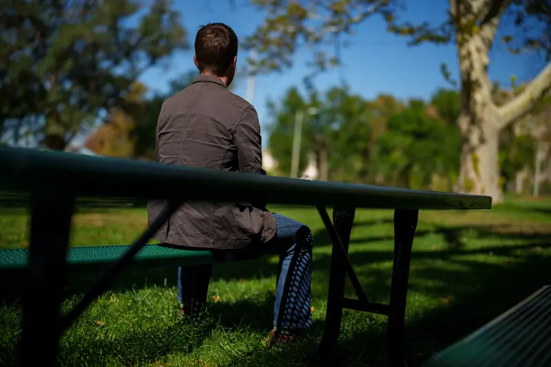 A brown-haired man sits on a park bench, facing away from the camera.